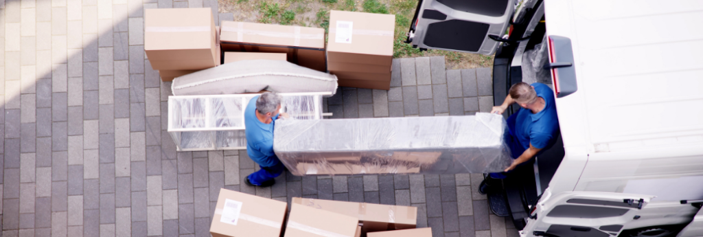 Two removalists carrying wrapped furniture from a van past stacked boxes